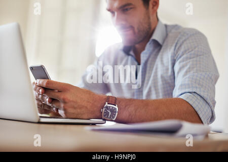 Shot of young businessman using mobile phone at home. L'homme assis à table avec ordinateur portable et lire un message texte sur son téléphone intelligent. Banque D'Images