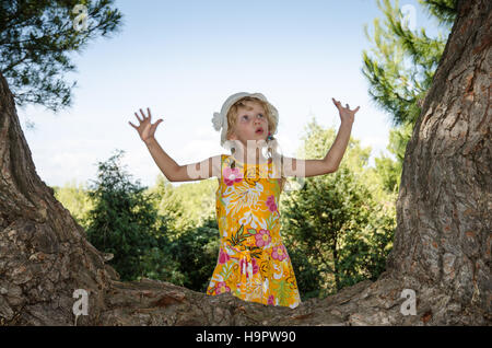 Belle blonde petite fille debout près du tronc de l'arbre avec surprise l'expression avec geste sorcière Banque D'Images