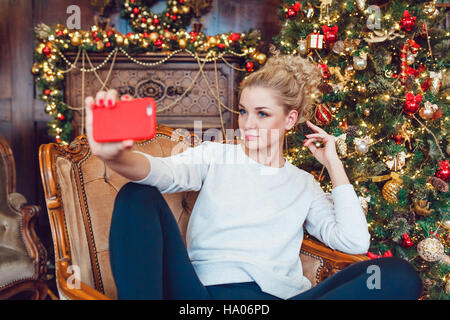 Très jolie jeune femme assise sur une chaise près de l'arbre de Noël et de faire. selfies Banque D'Images