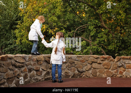 Cute Little Sisters dans le parc de l'automne. La fille plus âgée aider les jeunes rendez-vous sur la clôture en pierre. Le temps passé en famille. Jeux de sports. Banque D'Images