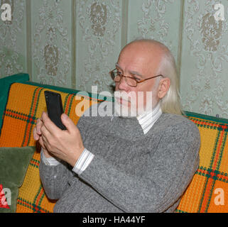 Portrait demi-longueur de pygargues à tête homme âgé avec de longs cheveux gris et sa barbe en gris chandail tricoté smartphone est assise sur le canapé. Banque D'Images