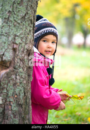 Kid in autumn park caché derrière tree et sourire Banque D'Images