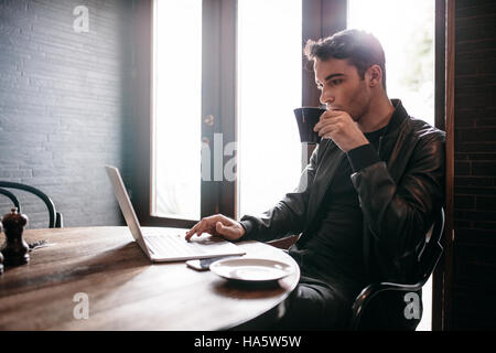 Tourné à l'intérieur du jeune homme assis à une table de café à boire du café et à l'aide d'un ordinateur portable. Banque D'Images