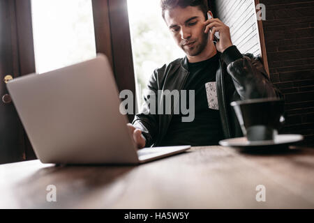 Photo de beau jeune homme talking on cell phone alors qu'il était assis dans un café avec un ordinateur portable Banque D'Images