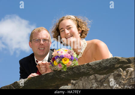 Mariée et le marié s'appuyant sur un vieux mur de château, tenant un bouquet de fleurs Banque D'Images