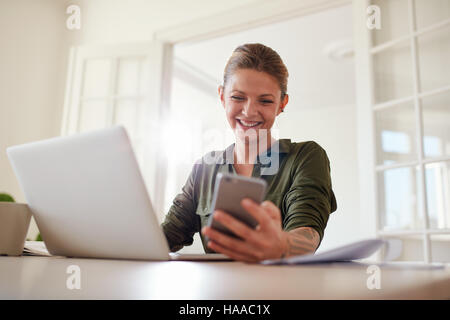 Shot de happy young woman using mobile phone while working on laptop. Femme de la lecture de texte message sur son téléphone intelligent et souriant. Banque D'Images