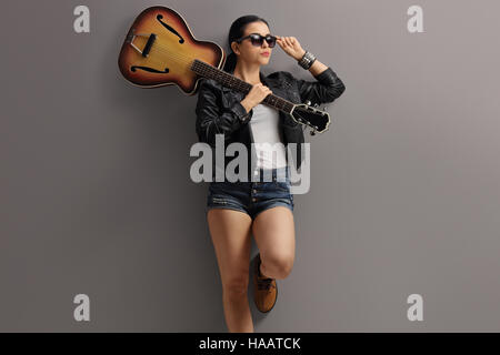 Femme dans un manteau de cuir qui posent avec une guitare et appuyé contre un mur gris Banque D'Images