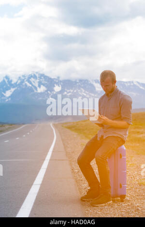 Seul l'homme est assis sur une valise et à la recherche à tablette. En attente d'un bus ou une voiture. La route dans les montagnes. Banque D'Images