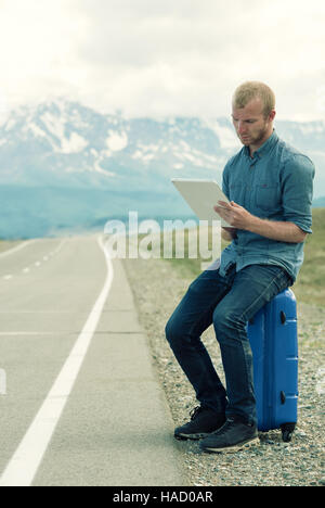 Seul l'homme est assis sur une valise et à la recherche à tablette. En attente d'un bus ou une voiture. La route dans les montagnes. Banque D'Images