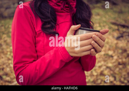 Dame avec pull rouge tenant dans ses mains le thé chaud dans la forêt à l'automne avec beaucoup de feuilles jaunes et arbres en arrière-plan.. Banque D'Images