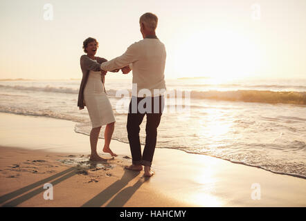 Toute la longueur de balle happy senior couple having fun sur la plage. Couple au bord de la mer se tenant la main au coucher du soleil. Banque D'Images