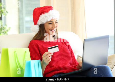 Woman wearing red sweater et santa hat l'achat des cadeaux de noël en ligne en utilisant un ordinateur portable assis sur un canapé dans la salle de séjour à la maison Banque D'Images