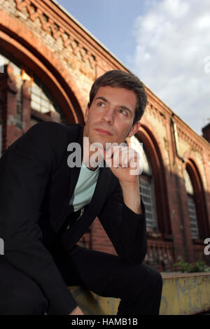 Portrait d'un jeune homme portant un costume, assis en face d'un bâtiment industriel en briques rouges à la recherche dans la distance Banque D'Images