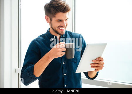 Photo de jeunes happy man holding tablet et boire du café près de la fenêtre. À la recherche de tablette. Banque D'Images