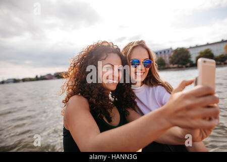 Shot of teenage girls smiling et en tenant avec selfies smart phone par le lac. Female friends taking self portrait avec téléphone mobile. Banque D'Images