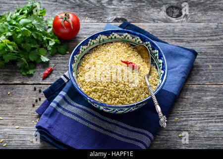 Les grains de blé bulgur brutes dans l'arabe coloré bol, du persil frais, tomates et poivrons pour la cuisson. Arrière-plan de table en bois Banque D'Images