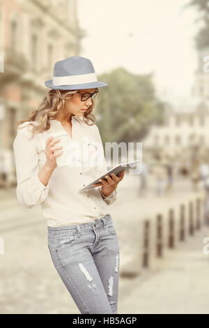 Belle jeune femme avec une tasse à café jetable, holding tablet dans ses mains, la lecture des nouvelles et des études urbaines contre l'arrière-plan de la ville. Banque D'Images