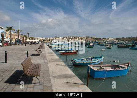 Vue sur le port de pêche de Marsaxlokk à Malte montrant la promenade, les bâtiments et les petits bateaux de pêche contre un ciel bleu Banque D'Images