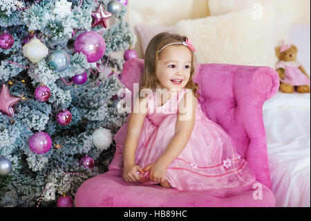 Happy happy little girl excité à la veille de Noël, assis sous l'arbre lumineux décoré. Carte de vœux ou couvrir, avec copie espace horizontal. Banque D'Images