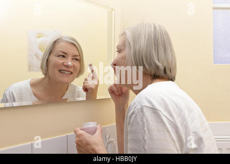 Senior woman with cream pot dans la main et une tache blanche sur le nez devant un miroir de salle de bains jaune applying moisturizer Banque D'Images