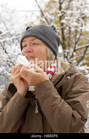 Hauts femme avec un nez rouge en hiver, manteau, écharpe et hat éternuer dans un mouchoir en papier en face d'un arbre avec de la neige Banque D'Images