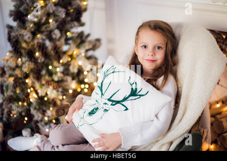 Fille à Noël, arbre de Noël en arrière-plan, jeune et belle enfant enveloppé dans une couverture, elle sourit Banque D'Images