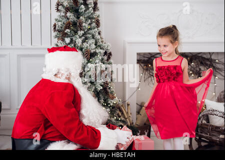 Le Père Noël et les enfants présents à l'ouverture de cheminée. Enfants père en costume portant barbe ouvrir les cadeaux de Noël. Petite fille aidant avec sac actuel. En vertu de la famille arbre de Noël plus de cheminée sur l'arrière-plan. Banque D'Images