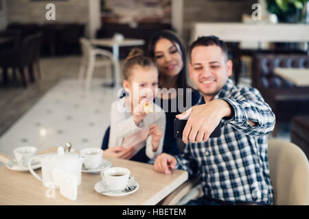 La famille, la parentalité, la technologie les gens heureux concept - mère, père et petite fille ayant pris le dîner au restaurant du smartphone par selfies Banque D'Images