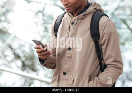 Image coupée de jeune homme teint foncé avec sac à dos chat par son téléphone dans la rue. Banque D'Images