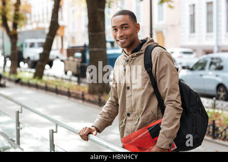 Photo de young African man wearing backpack debout dans la rue tout en maintenant réserver et looking at camera. Banque D'Images