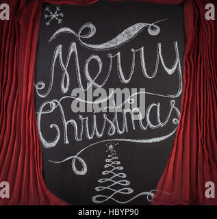 Joyeux Noël lettrage dessiné à la main avec de la craie sur tableau noir encadré par des rideaux rouges. Élégant design unique avec des flocons de neige et l'arbre de Noël Banque D'Images