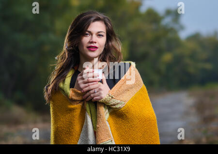 Belle jeune fille avec une tasse de café (thé) avec une couverture sur ses épaules à l'extérieur en soirée d'automne. Banque D'Images