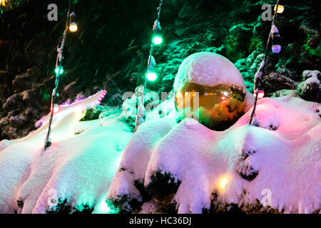 Grosse Boule de Noël avec de la neige dessus on snowy fir et les branches d'arbres entre les lumières de Noël coloré Banque D'Images