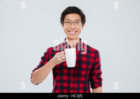 Photo de jeune homme habillé en chemise décontractée dans une cage et portant des lunettes holding tasse de thé. Plus isolé sur fond blanc. À la recherche à Banque D'Images