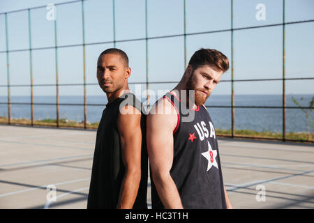 Deux jeunes sportifs miltiracial debout dos à dos à l'aire de jeux et looking at camera Banque D'Images