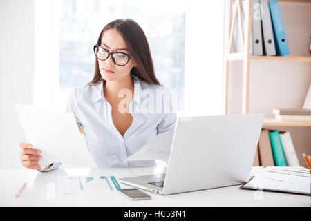 Photo de portrait habillé en chemise blanche et portant des lunettes de bureau et ordinateur portable à l'aide pendant la recherche de documents. Banque D'Images