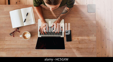Top View of woman typing on laptop mains. Femme assise à table avec café, ordinateur portable téléphone mobile, journal, tasse à café et verres. Banque D'Images