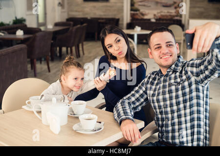 La famille, la parentalité, la technologie les gens heureux concept - mère, père et petite fille ayant pris le dîner au restaurant du smartphone par selfies Banque D'Images