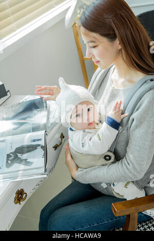 Vue latérale du smiling woman hugging her baby reading magazine Banque D'Images
