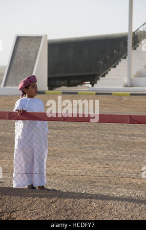 Un garçon en robe blanche et turban à l'hippodrome de chameau en Oman en attente les gagnants. Banque D'Images