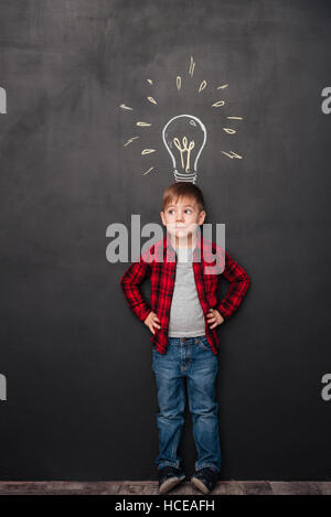 Photo d'un petit garçon mignon ayant une idée sur tableau arrière-plan avec des dessins. Regarder de côté. Banque D'Images