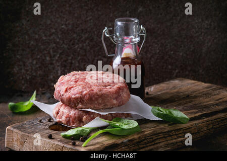 Ingrédients pour faire de hamburger. Escalope de boeuf hamburger cru, deux petits pains, oignon frit, épinards frais et sauce ketchup sur planche sombre plus vieux woode Banque D'Images