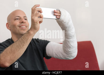Jeune homme avec un bras et coude dans un plâtre blanc / plâtre en fibre de verre à la maison, parler d'un sur son téléphone intelligent selfies lors de la récupération d'un bras cassé Banque D'Images