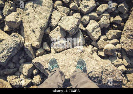 Les pieds mâles dans les chaussures de sport en toile bleu debout sur les zones côtières un sol rocailleux. Billet d'lifstyle arrière-plan. Correction tonale chaud vintage photo filtre, o Banque D'Images
