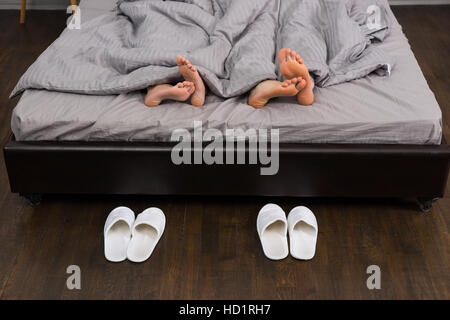 Les pieds mâles et femelles sous couverture grise au lit moderne et élégant près de deux paires de chaussons blanc Banque D'Images