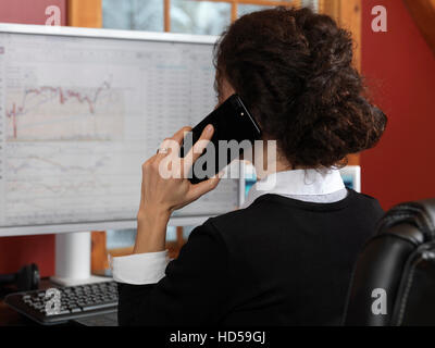 Femme parlant stock trader sur le téléphone portable Apple iPhone 7 plus graphiques boursiers avec affiché sur son écran d'ordinateur Banque D'Images