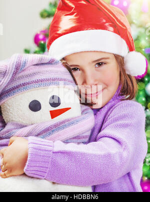 Closeup portrait of a cute little baby girl wearing Santa hat rouge près de l'arbre de Noël la décoration de fête, avec l'amour l'étreindre soft toy snowman Banque D'Images