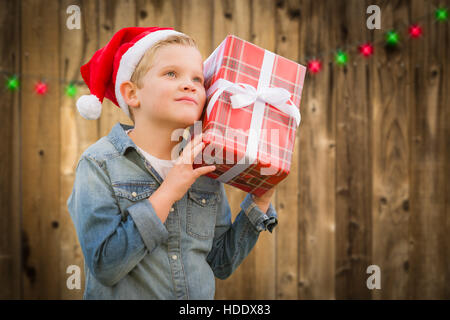 Jeune garçon curieux Wearing Santa Hat Holding Christmas Gift sur une barrière en bois. Banque D'Images