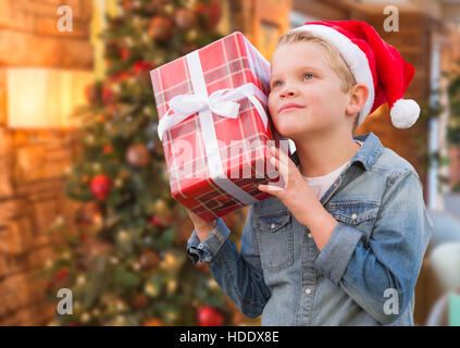 Jeune garçon curieux Wearing Santa Hat Holding Christmas Gift en face d'un arbre décoré. Banque D'Images