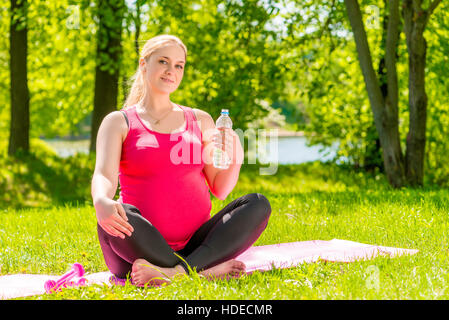 Jeune femme enceinte a un reste assis sur un tapis avec une bouteille d'eau dans le parc Banque D'Images